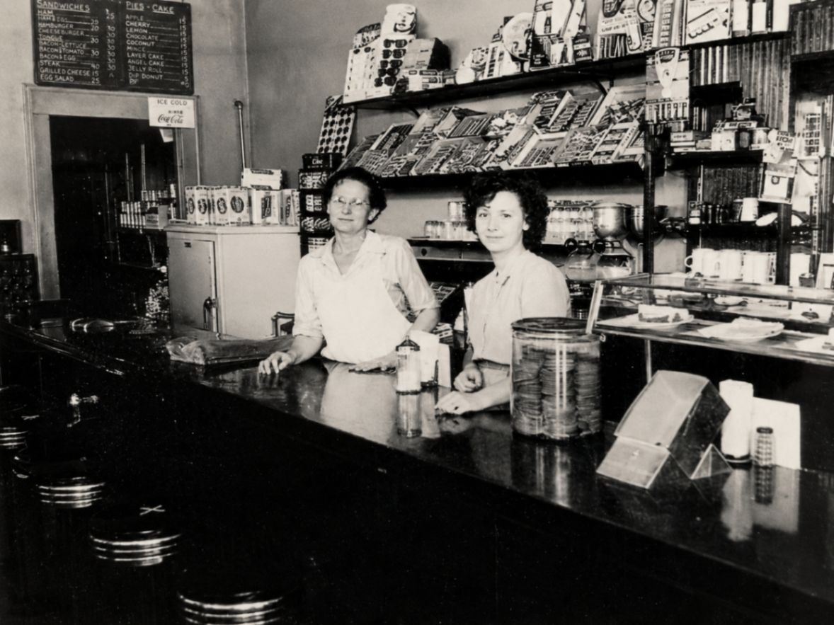Two women behind a soda fountain counter with a sandwich and pie menu board